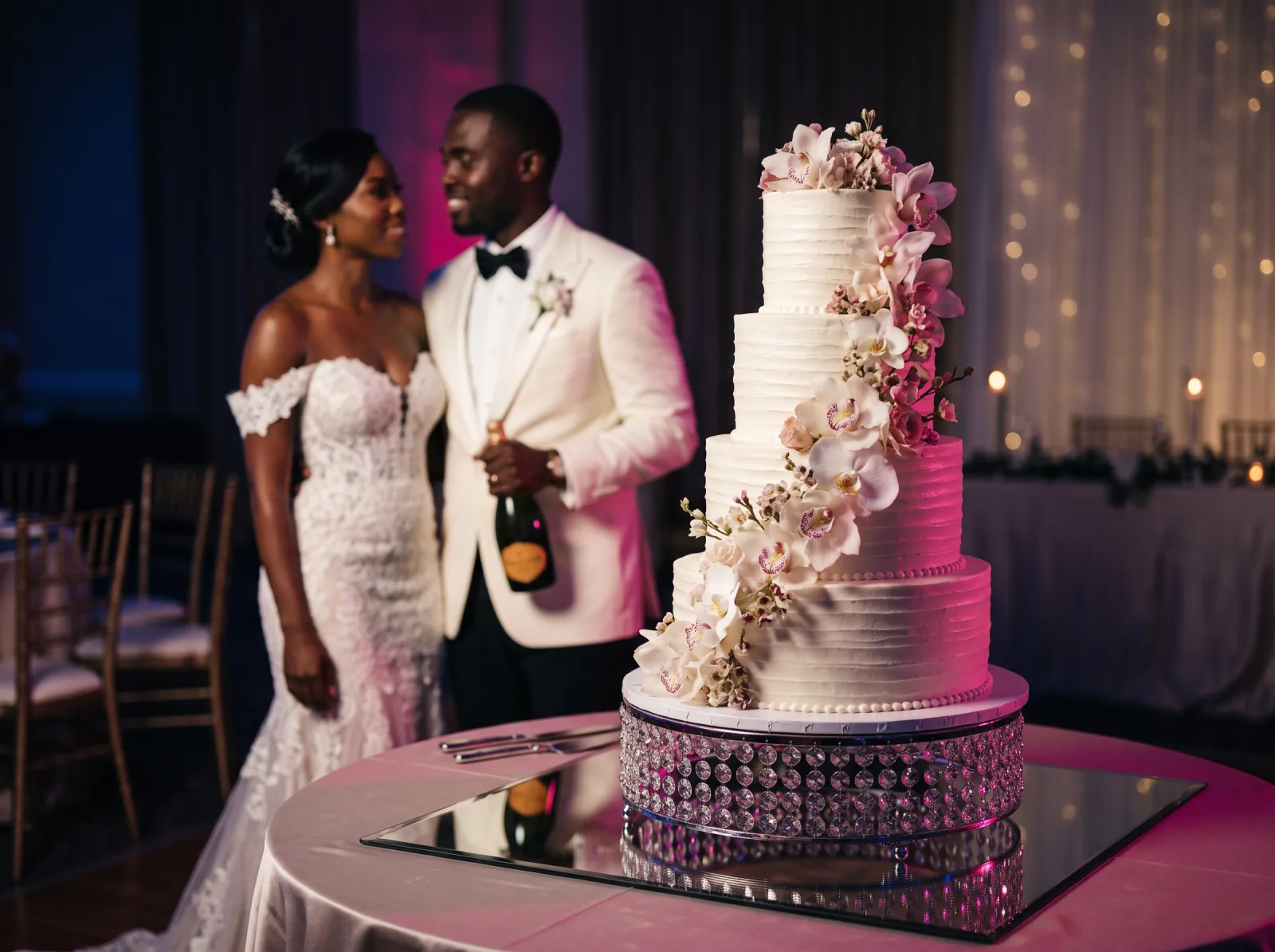 Bride and groom standing beside a tall uncut tiered wedding cake trimmed with orchids at a Haitian reception